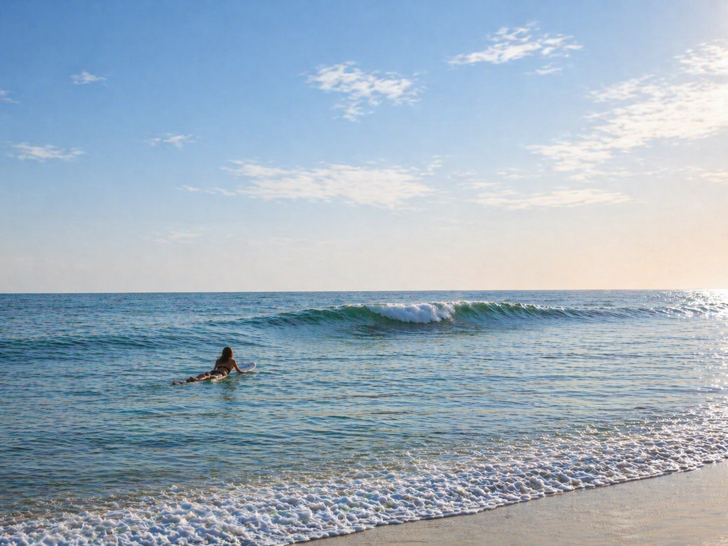 A woman paddling into a small, glassy wave, sunlight rim-lighting her silhouette.